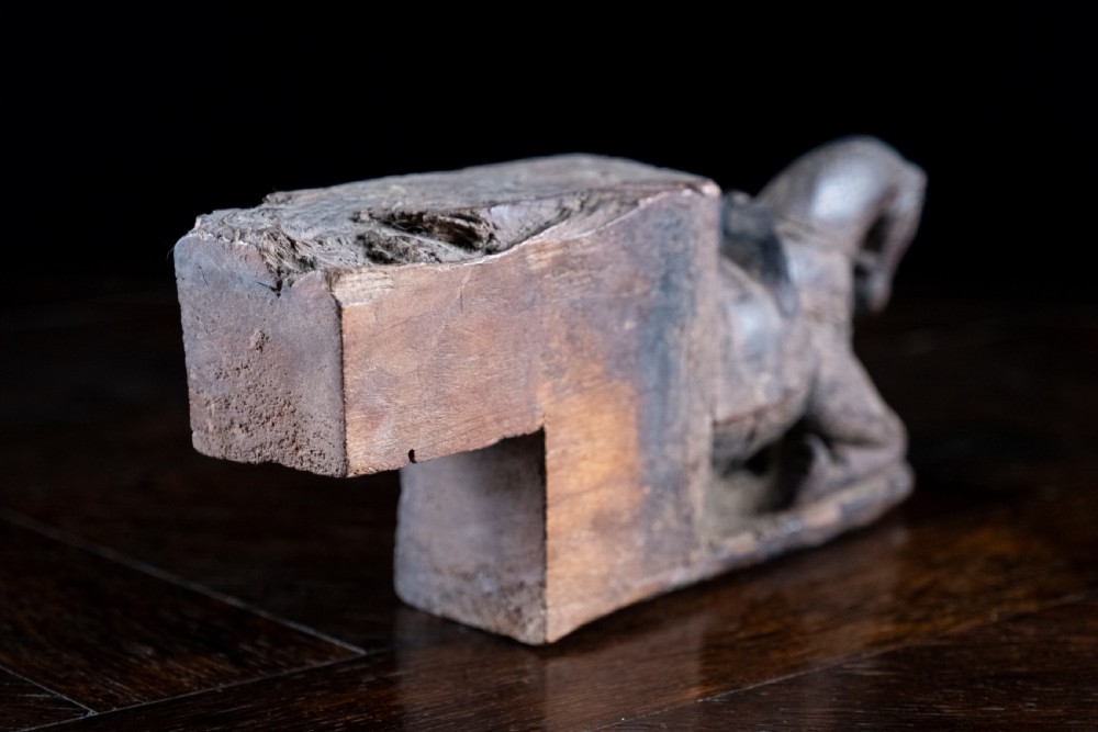 A close-up photograph of a weathered, dark brown carved wooden horse architectural element shown from a three-quarters front angle against a black background. View VII