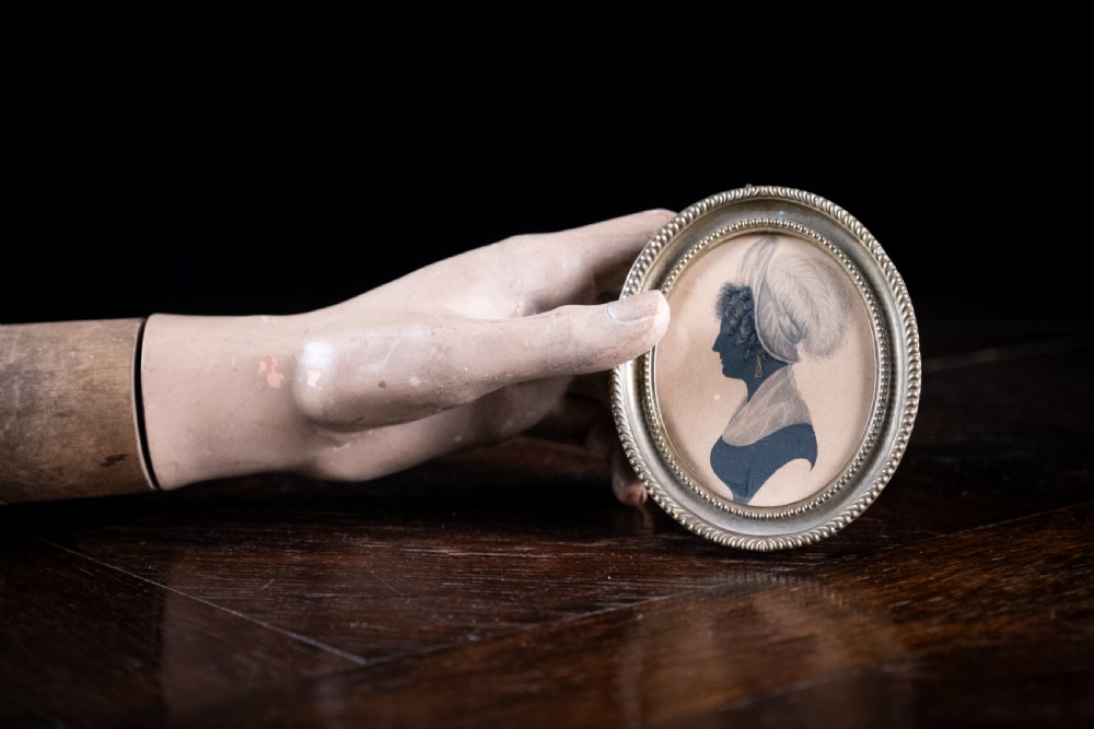 An oval early nineteenth-century silhouette portrait of a woman in profile featuring gilded details, housed in a beaded metal frame. View IV