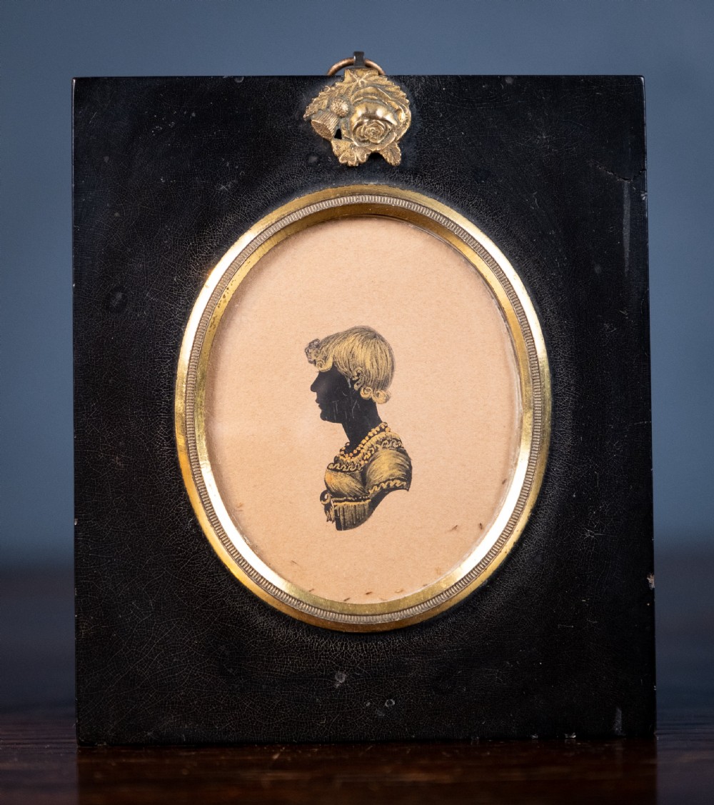 A small oval silhouette portrait of a profile with gilded details, enclosed in a brass oval bezel inside a black rectangular papier-mache frame with a brass floral hanger.