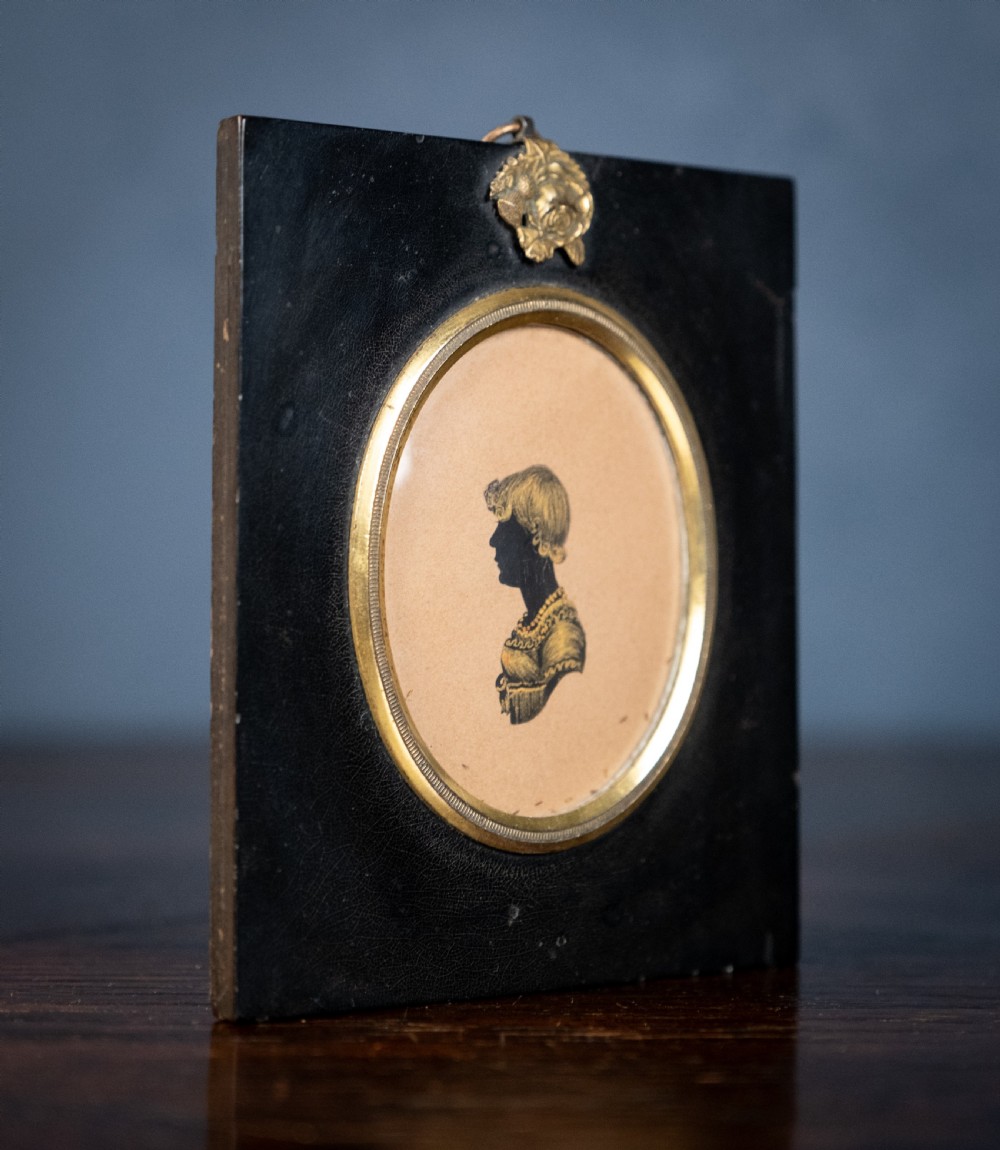 A small oval silhouette portrait of a profile with gilded details, enclosed in a brass oval bezel inside a black rectangular papier-mache frame with a brass floral hanger. View III