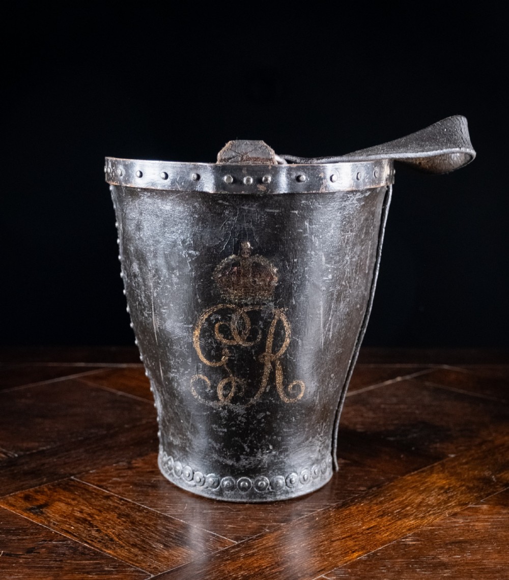 A black leather fire bucket featuring a royal cipher and reinforced metal rim, positioned on a wooden surface against a dark background.