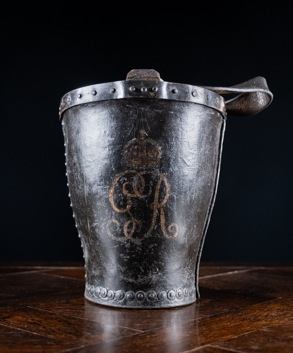 A black leather fire bucket featuring a royal cipher and reinforced metal rim, positioned on a wooden surface against a dark background. View II
