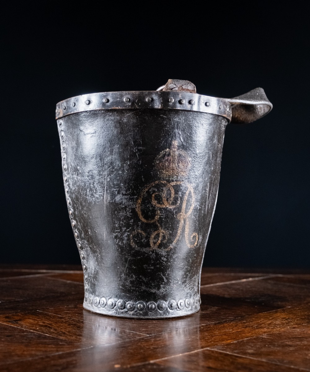 A black leather fire bucket featuring a royal cipher and reinforced metal rim, positioned on a wooden surface against a dark background. View III