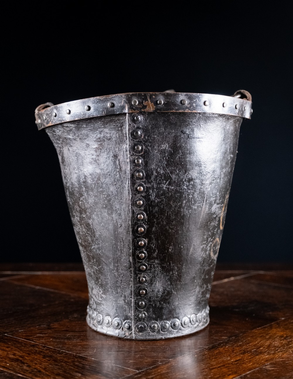 A black leather fire bucket featuring a royal cipher and reinforced metal rim, positioned on a wooden surface against a dark background. View IV