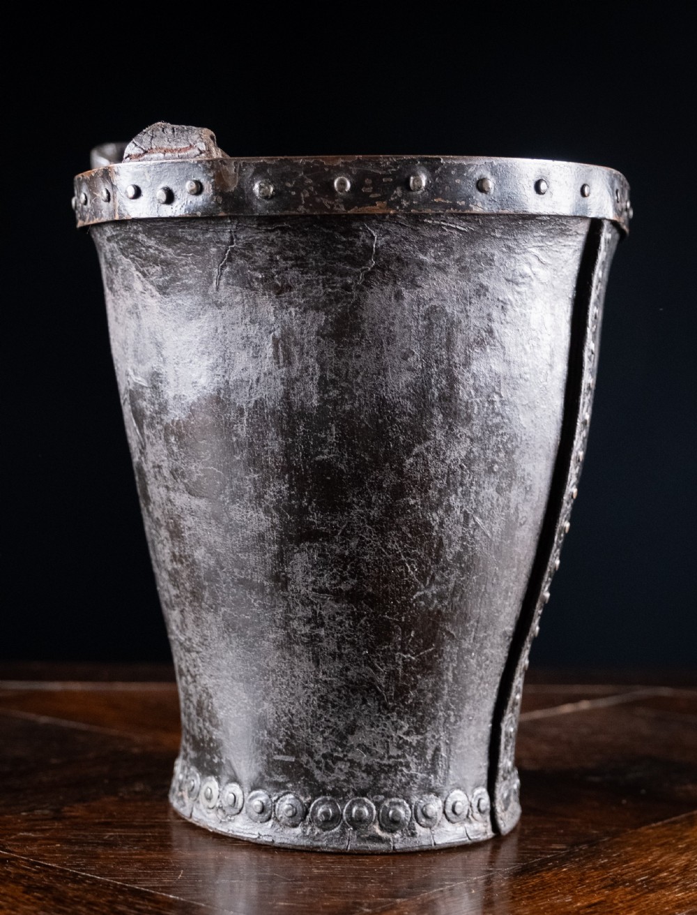 A black leather fire bucket featuring a royal cipher and reinforced metal rim, positioned on a wooden surface against a dark background. View V