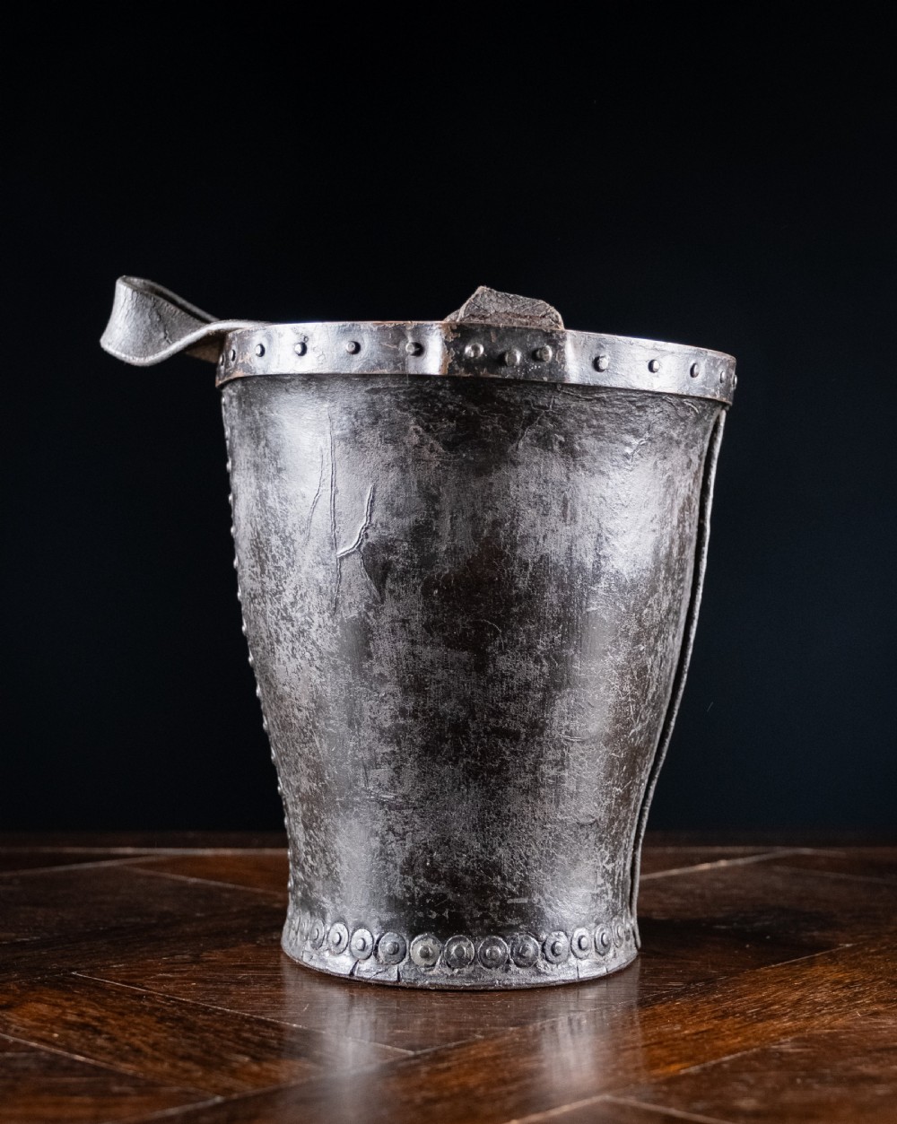 A black leather fire bucket featuring a royal cipher and reinforced metal rim, positioned on a wooden surface against a dark background. View VI