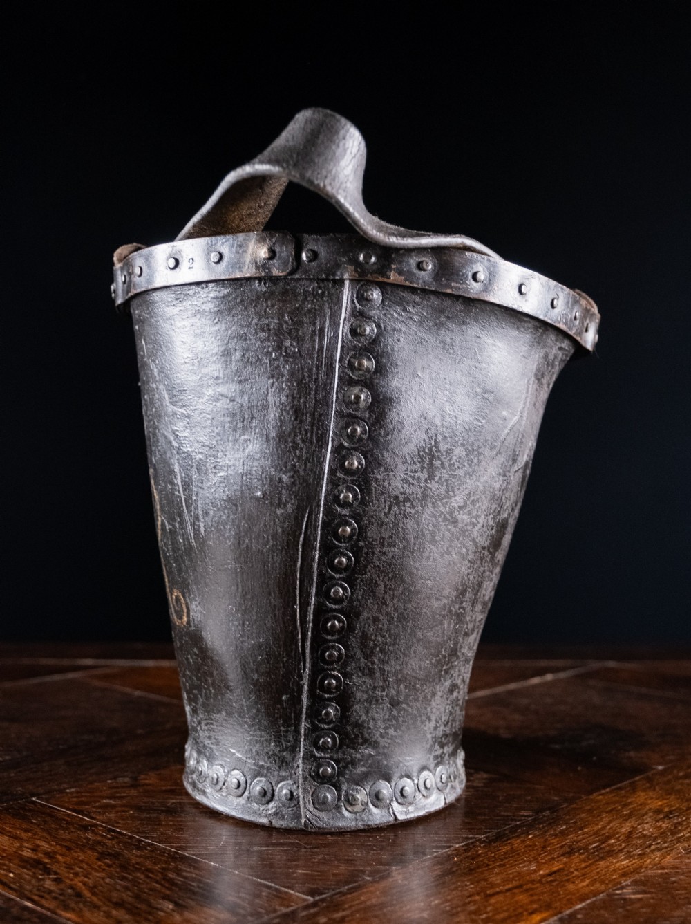 A black leather fire bucket featuring a royal cipher and reinforced metal rim, positioned on a wooden surface against a dark background. View VII