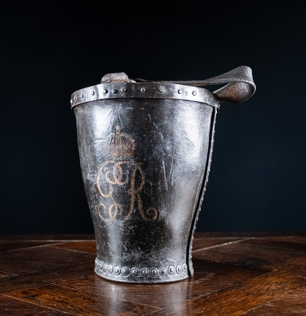 A black leather fire bucket featuring a royal cipher and reinforced metal rim, positioned on a wooden surface against a dark background. View VIII