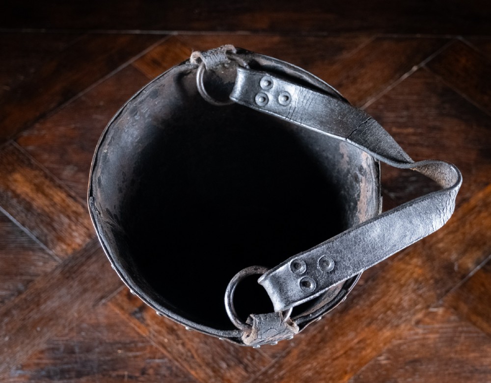 A black leather fire bucket featuring a royal cipher and reinforced metal rim, positioned on a wooden surface against a dark background. View IX