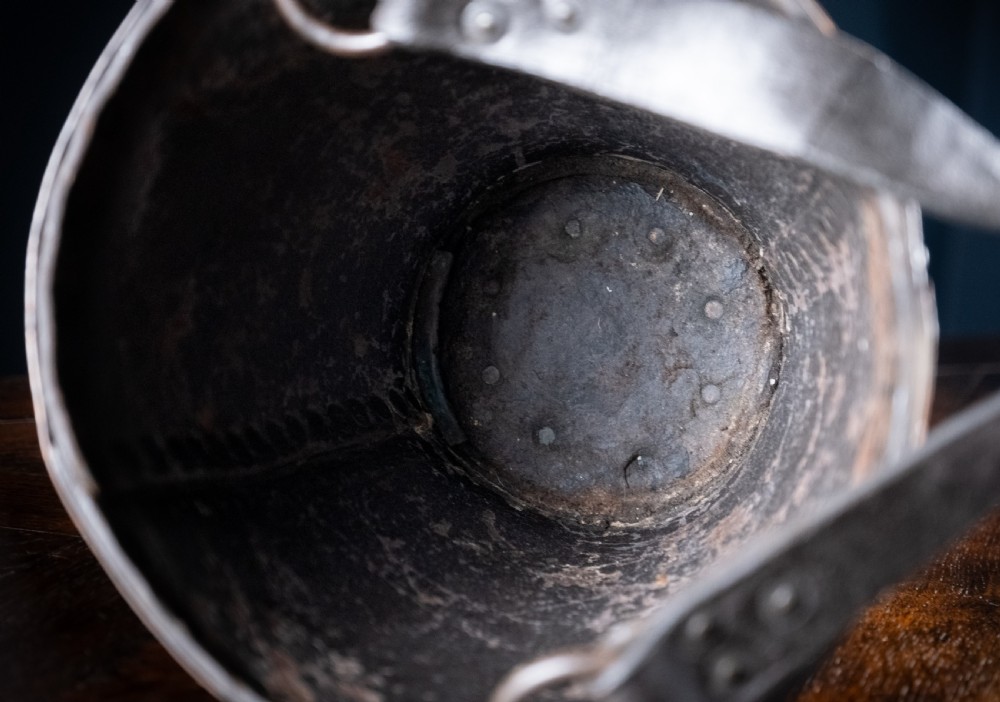 A black leather fire bucket featuring a royal cipher and reinforced metal rim, positioned on a wooden surface against a dark background. View X
