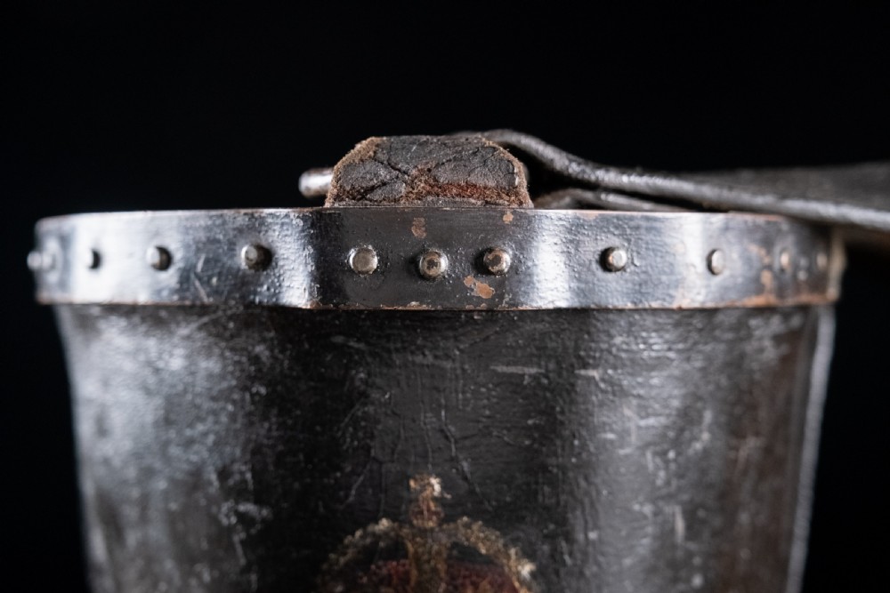 A black leather fire bucket featuring a royal cipher and reinforced metal rim, positioned on a wooden surface against a dark background. View XII