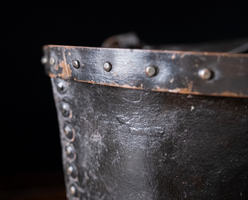A black leather fire bucket featuring a royal cipher and reinforced metal rim, positioned on a wooden surface against a dark background. View XIII