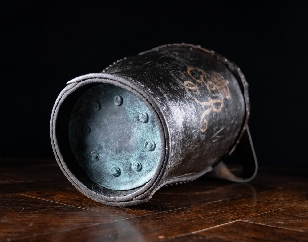 A black leather fire bucket featuring a royal cipher and reinforced metal rim, positioned on a wooden surface against a dark background. View XIV