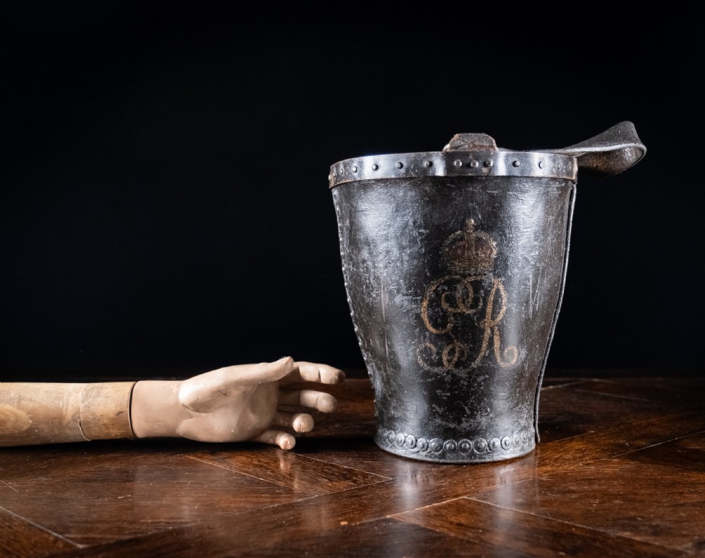 A black leather fire bucket featuring a royal cipher and reinforced metal rim, positioned on a wooden surface against a dark background. View XV