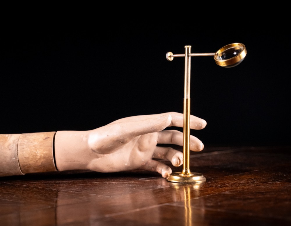 A lacquered brass bullseye condenser magnifying glass featuring an adjustable stand and a convex lens, positioned on a dark wooden surface.