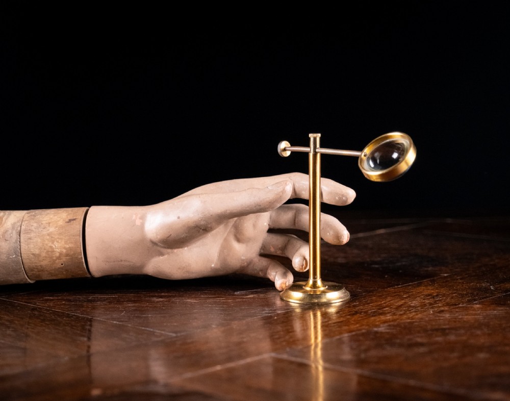 A lacquered brass bullseye condenser magnifying glass featuring an adjustable stand and a convex lens, positioned on a dark wooden surface. View II