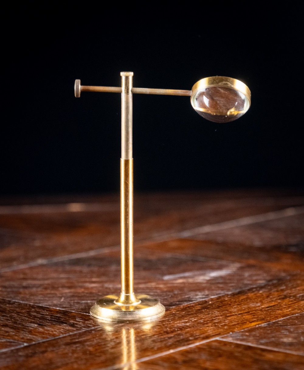 A lacquered brass bullseye condenser magnifying glass featuring an adjustable stand and a convex lens, positioned on a dark wooden surface. View III