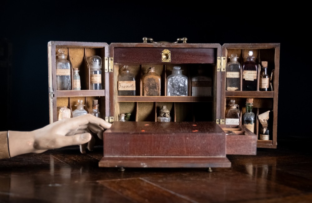 An open antique wooden apothecary cabinet featuring multiple compartments filled with vintage glass medicine bottles. View III
