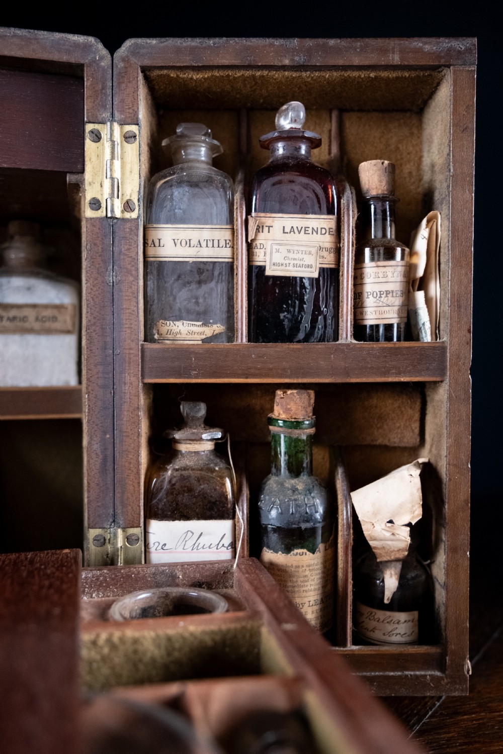 An open antique wooden apothecary cabinet featuring multiple compartments filled with vintage glass medicine bottles. View V