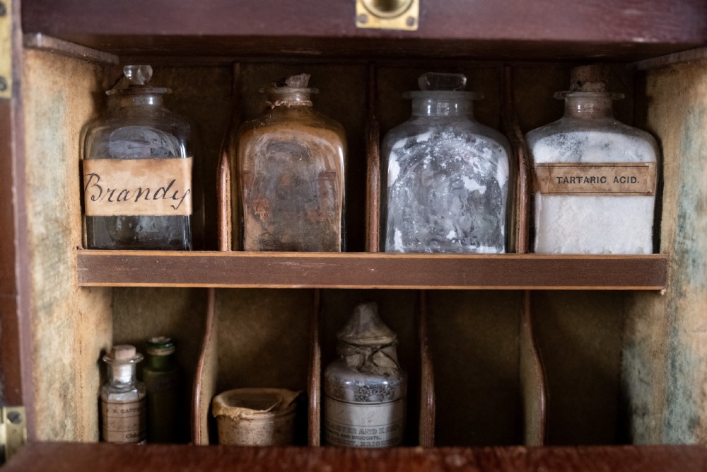 An open antique wooden apothecary cabinet featuring multiple compartments filled with vintage glass medicine bottles. View VI