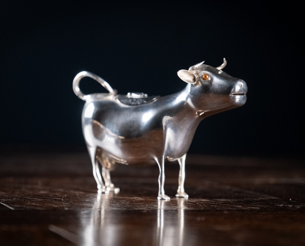 A small silver cow creamer standing on four slender legs with a hinged lid on its back and a looped tail acting as a handle.