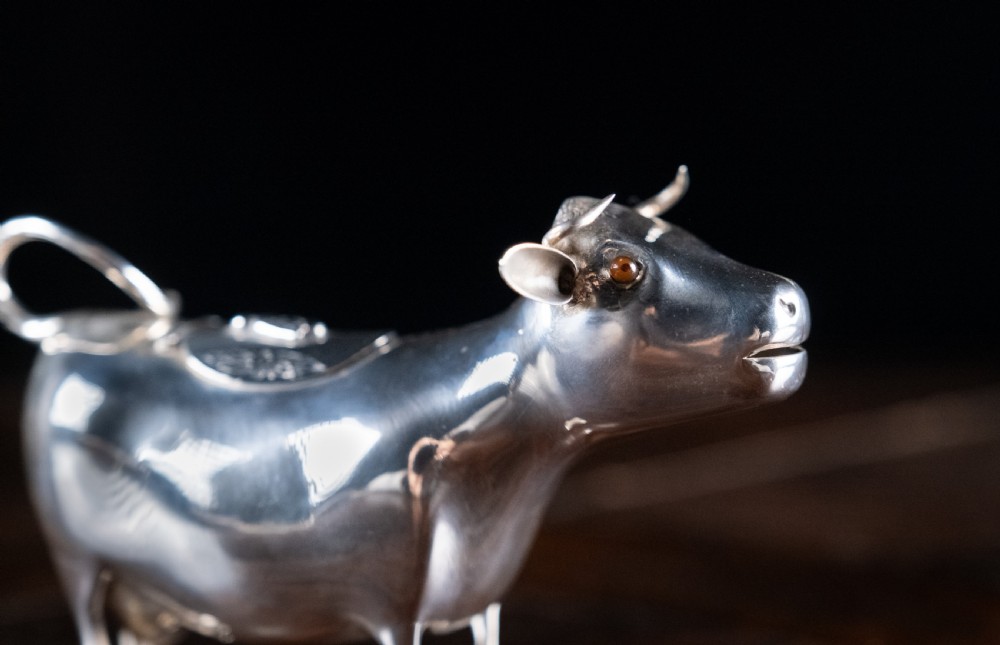 A small silver cow creamer standing on four slender legs with a hinged lid on its back and a looped tail acting as a handle. View II
