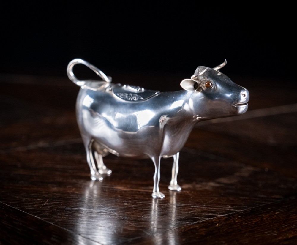 A small silver cow creamer standing on four slender legs with a hinged lid on its back and a looped tail acting as a handle. View IV