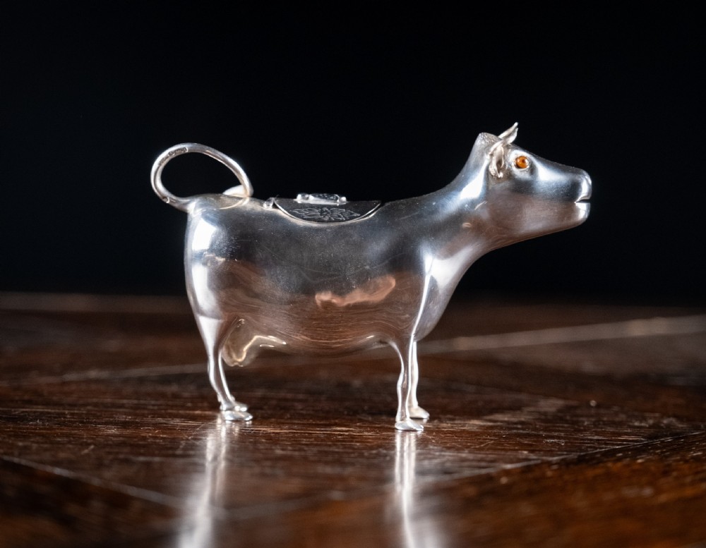 A small silver cow creamer standing on four slender legs with a hinged lid on its back and a looped tail acting as a handle. View VI