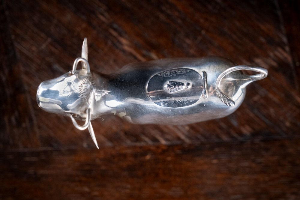 A small silver cow creamer standing on four slender legs with a hinged lid on its back and a looped tail acting as a handle. View X