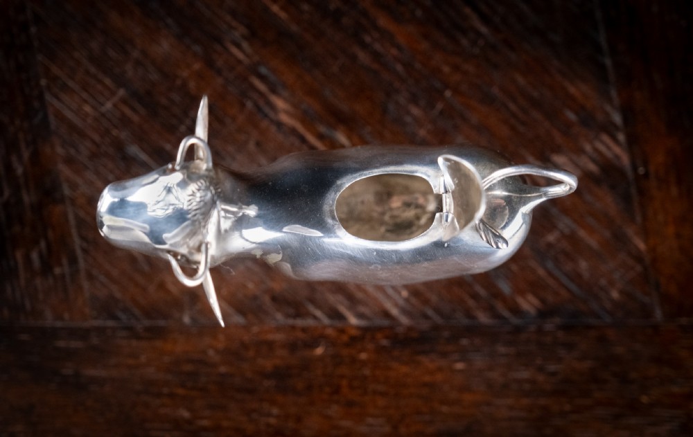 A small silver cow creamer standing on four slender legs with a hinged lid on its back and a looped tail acting as a handle. View XI
