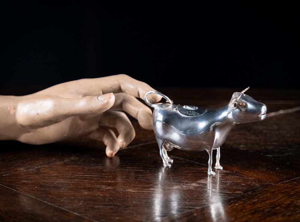 A small silver cow creamer standing on four slender legs with a hinged lid on its back and a looped tail acting as a handle. View XIII