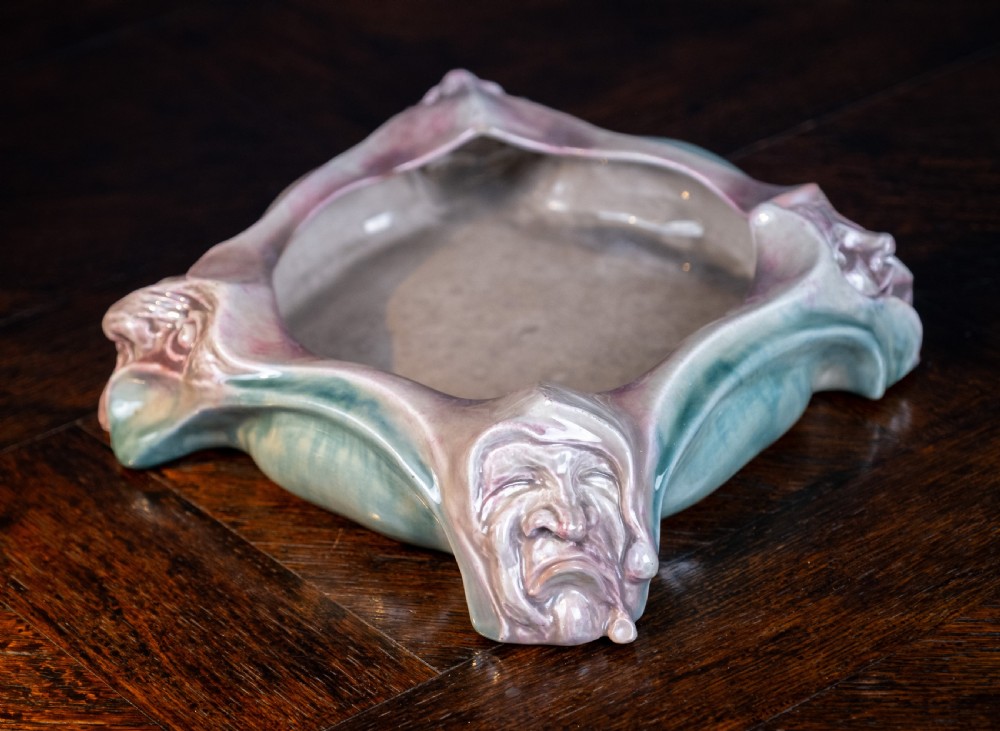 A square-shaped Art Nouveau glazed ceramic centre bowl featuring sculpted facial masks at each corner in soft green and iridescent pink glazes.
