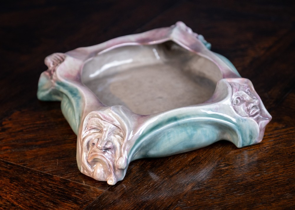 A square-shaped Art Nouveau glazed ceramic centre bowl featuring sculpted facial masks at each corner in soft green and iridescent pink glazes. View II