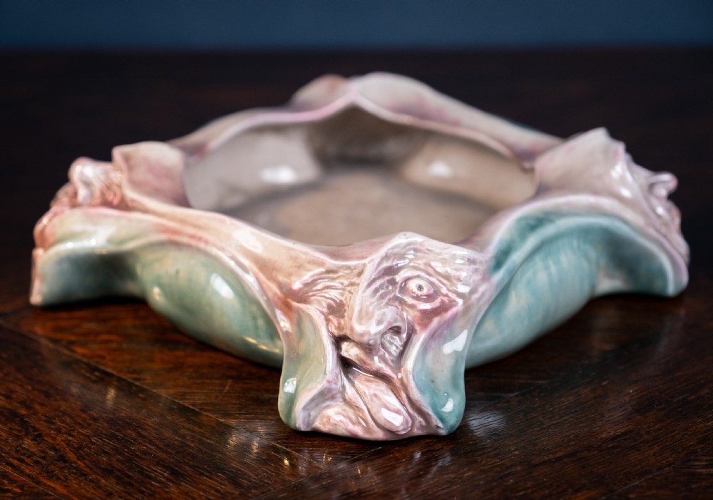 A square-shaped Art Nouveau glazed ceramic centre bowl featuring sculpted facial masks at each corner in soft green and iridescent pink glazes. View III