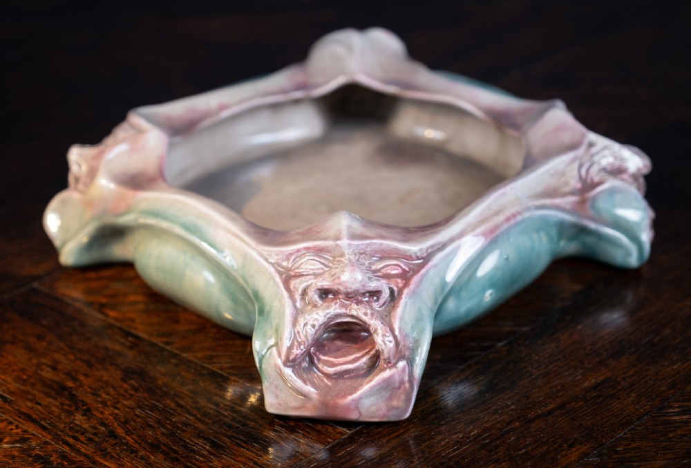 A square-shaped Art Nouveau glazed ceramic centre bowl featuring sculpted facial masks at each corner in soft green and iridescent pink glazes. View V
