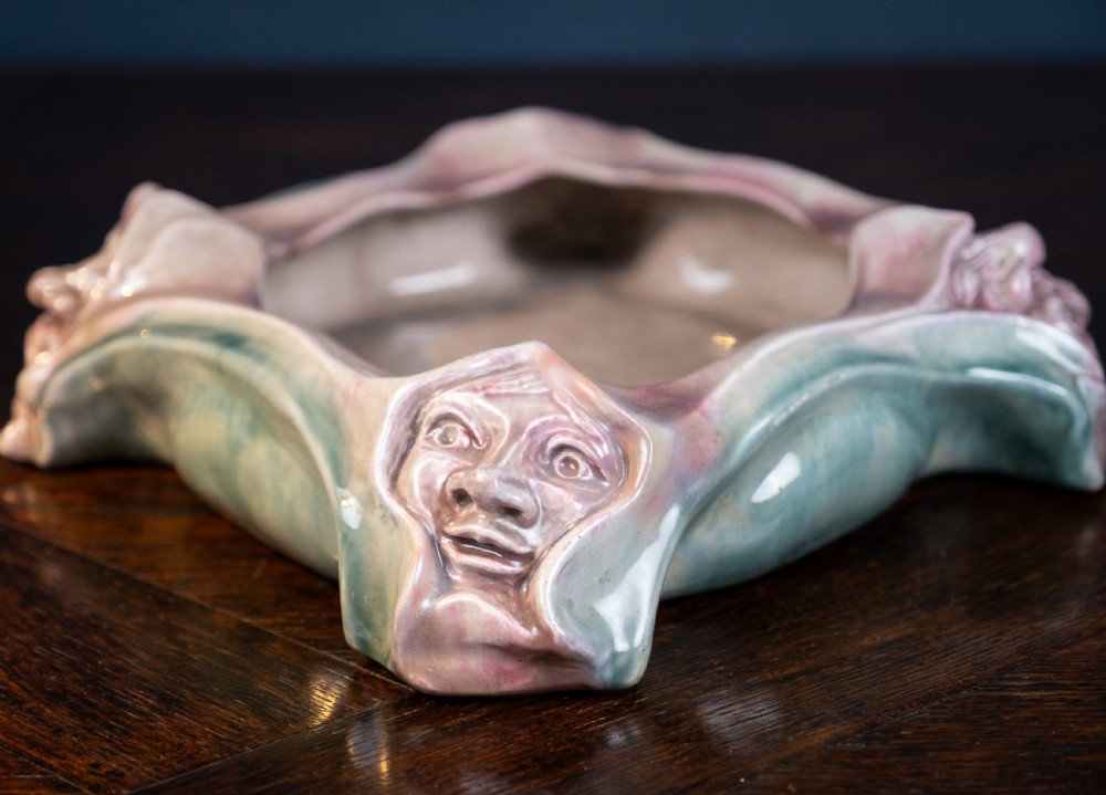 A square-shaped Art Nouveau glazed ceramic centre bowl featuring sculpted facial masks at each corner in soft green and iridescent pink glazes. View VI