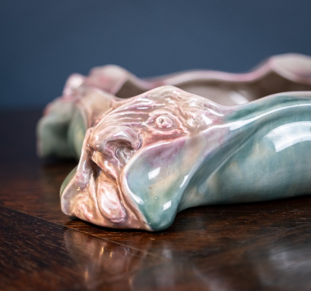A square-shaped Art Nouveau glazed ceramic centre bowl featuring sculpted facial masks at each corner in soft green and iridescent pink glazes. View VII