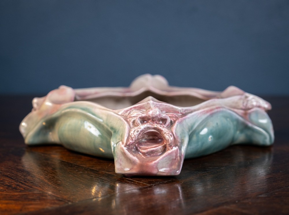 A square-shaped Art Nouveau glazed ceramic centre bowl featuring sculpted facial masks at each corner in soft green and iridescent pink glazes. View VIII