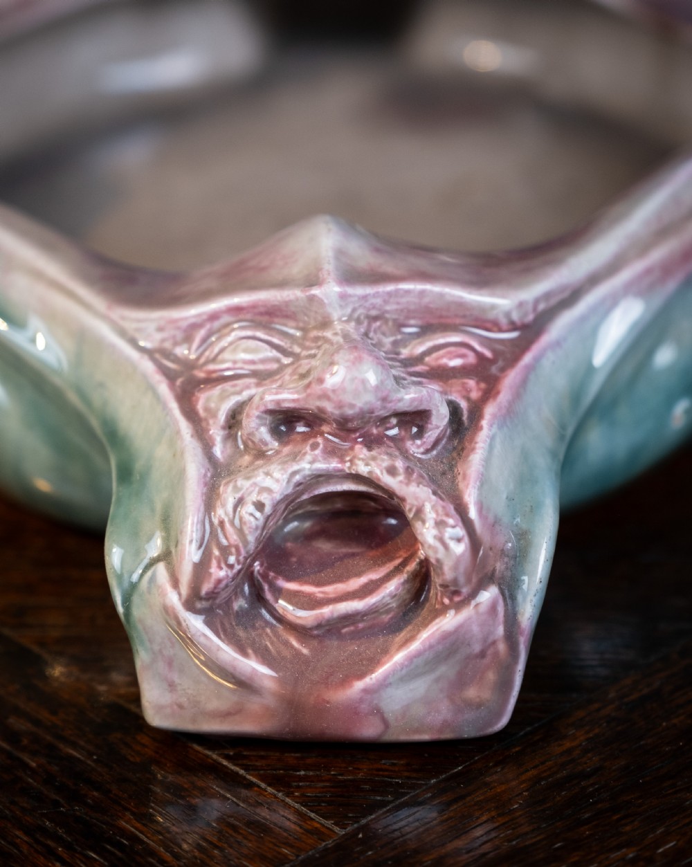 A square-shaped Art Nouveau glazed ceramic centre bowl featuring sculpted facial masks at each corner in soft green and iridescent pink glazes. View IX