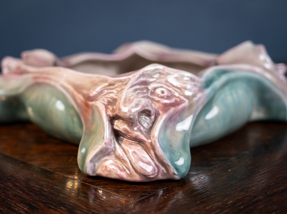 A square-shaped Art Nouveau glazed ceramic centre bowl featuring sculpted facial masks at each corner in soft green and iridescent pink glazes. View X