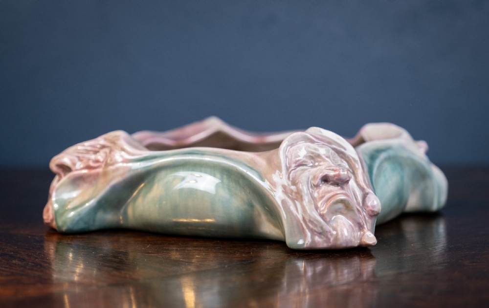 A square-shaped Art Nouveau glazed ceramic centre bowl featuring sculpted facial masks at each corner in soft green and iridescent pink glazes. View XI