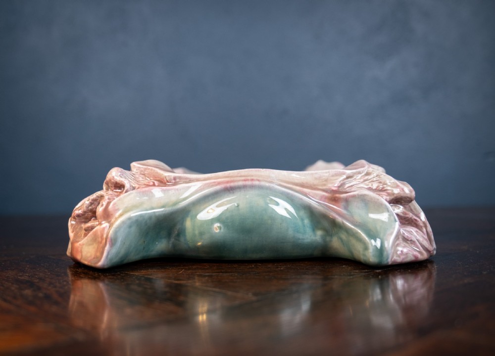 A square-shaped Art Nouveau glazed ceramic centre bowl featuring sculpted facial masks at each corner in soft green and iridescent pink glazes. View XII