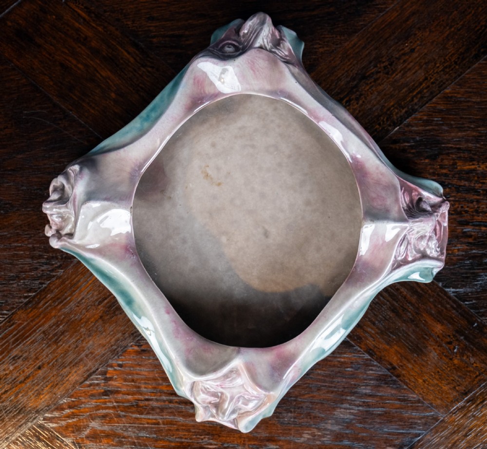 A square-shaped Art Nouveau glazed ceramic centre bowl featuring sculpted facial masks at each corner in soft green and iridescent pink glazes. View XIII