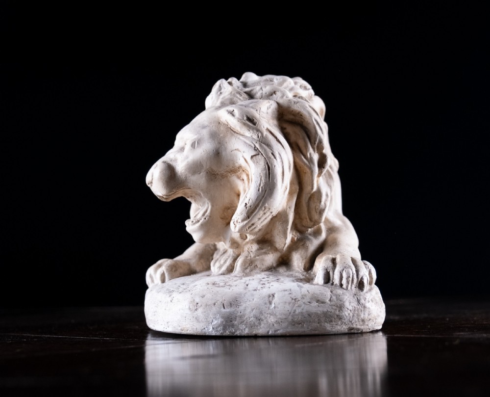 An off-white plaster sculpture depicting a prowling lion with an open mouth and detailed mane on an integral oval base. View V