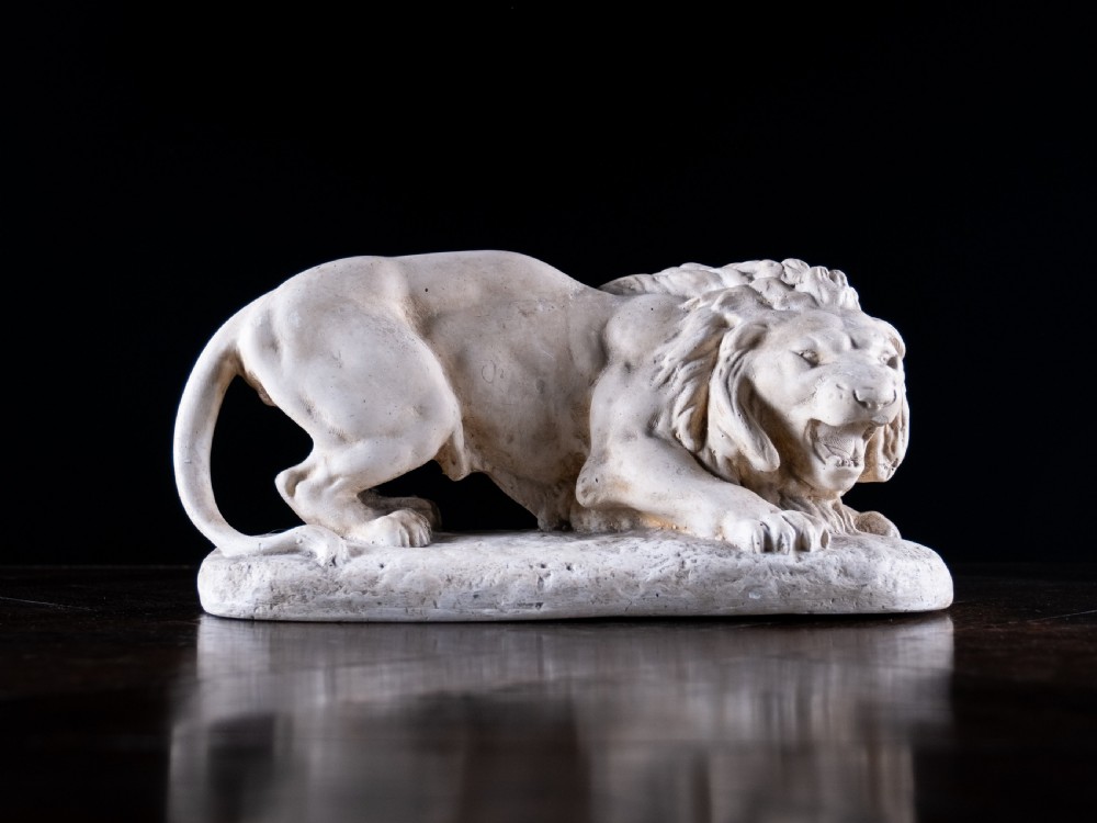 An off-white plaster sculpture depicting a prowling lion with an open mouth and detailed mane on an integral oval base. View X