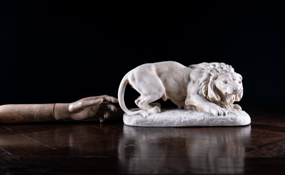 An off-white plaster sculpture depicting a prowling lion with an open mouth and detailed mane on an integral oval base. View XIII