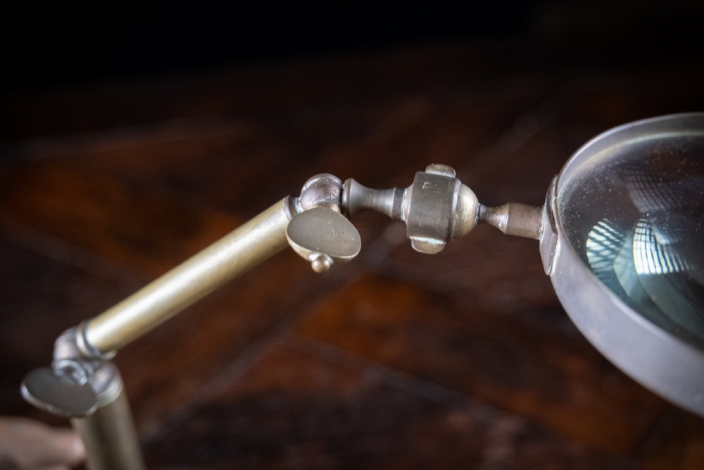 An antique Edwardian brass magnifying glass featuring an adjustable articulated arm and a circular weighted base. View VIII
