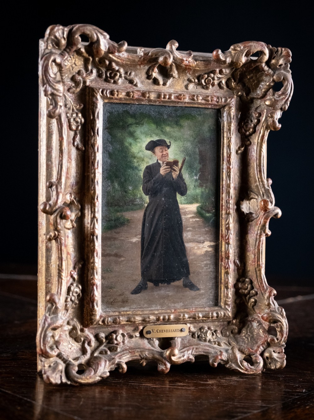 A 19th-century oil painting on panel depicting a smiling clergyman reading a book, set within a heavily carved rococo gilt frame. View II