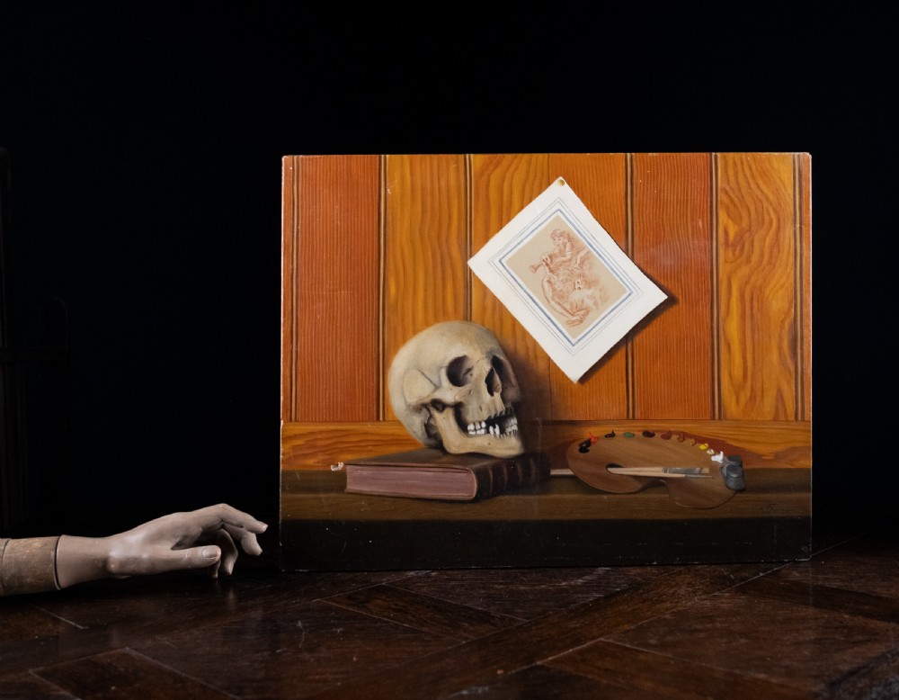 An oil painting depicting a human skull resting on a bound book alongside an artist palette, brushes, and a pinned sketch against a vertical timber panel background. View II