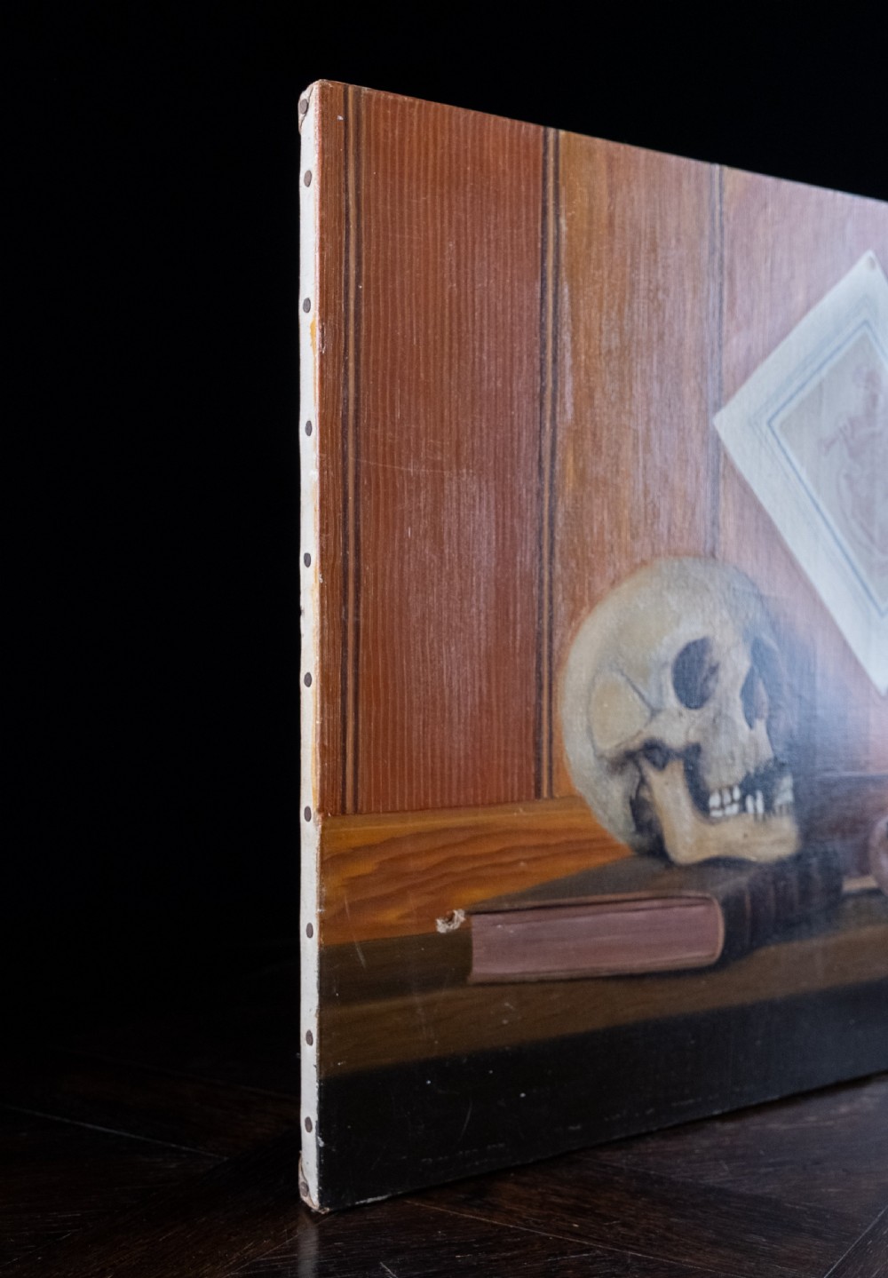 An oil painting depicting a human skull resting on a bound book alongside an artist palette, brushes, and a pinned sketch against a vertical timber panel background. View X